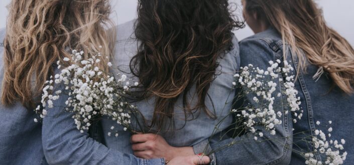 three women's backs with their hands holding dainty white flowers