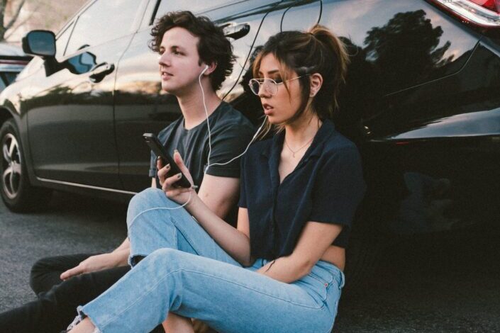 Couple, sitting against a car, listening to music together.