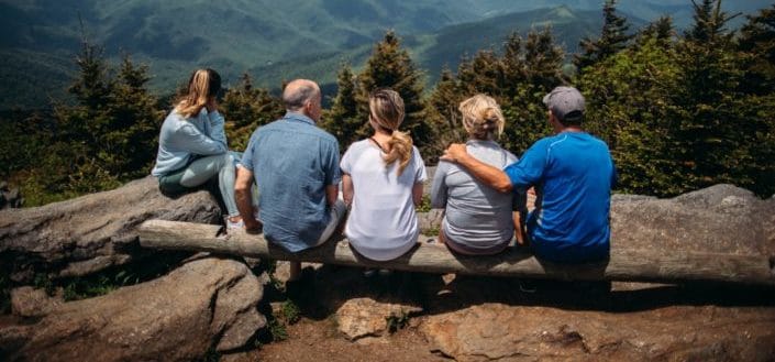 Group of elderly adults sitting on log in front of tall trees