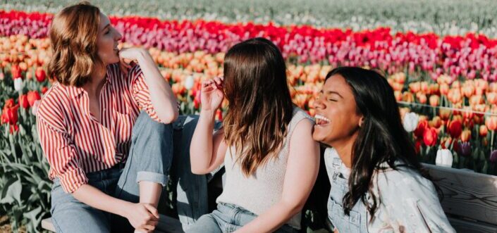 Three friends sitting and laughing on a bench in a flower garden.