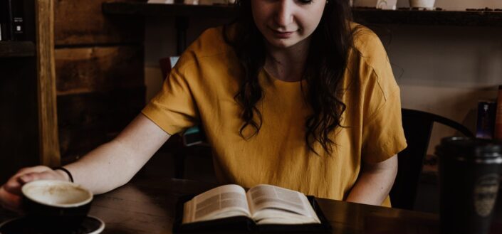 Woman sitting on a chair, reading at an open book laid on a table.