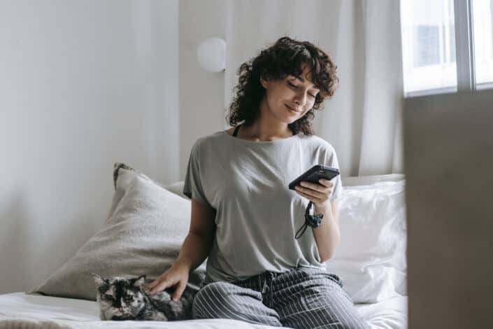 A woman with her cat beside her sitting on her bed smiling while looking at her phone