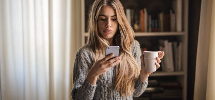 Woman in sweater looking at her phone while drinking coffee