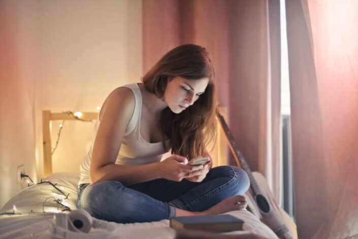 Photo of woman in white tank top and blue denim jeans sitting on bed while using her phone