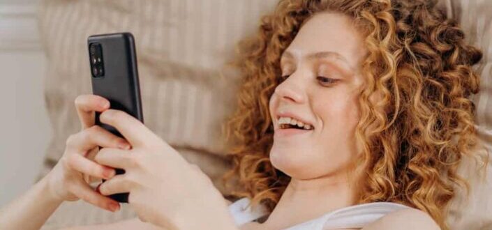 Woman in white tank top lying on bed holding black smartphone