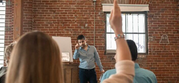 A bearded man pointing the pen at the person raising hand