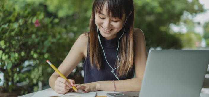 asian student writing while using laptop