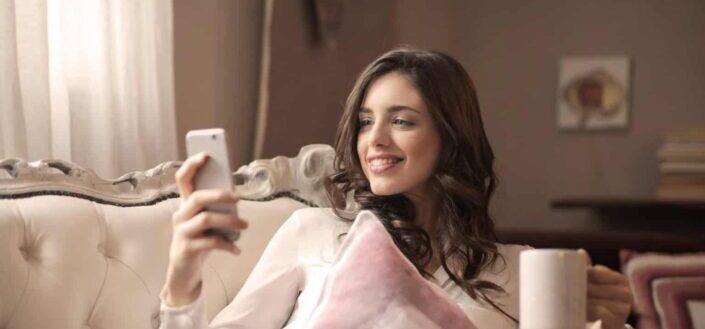 Woman Smiling at Phone Sitting On Sofa
